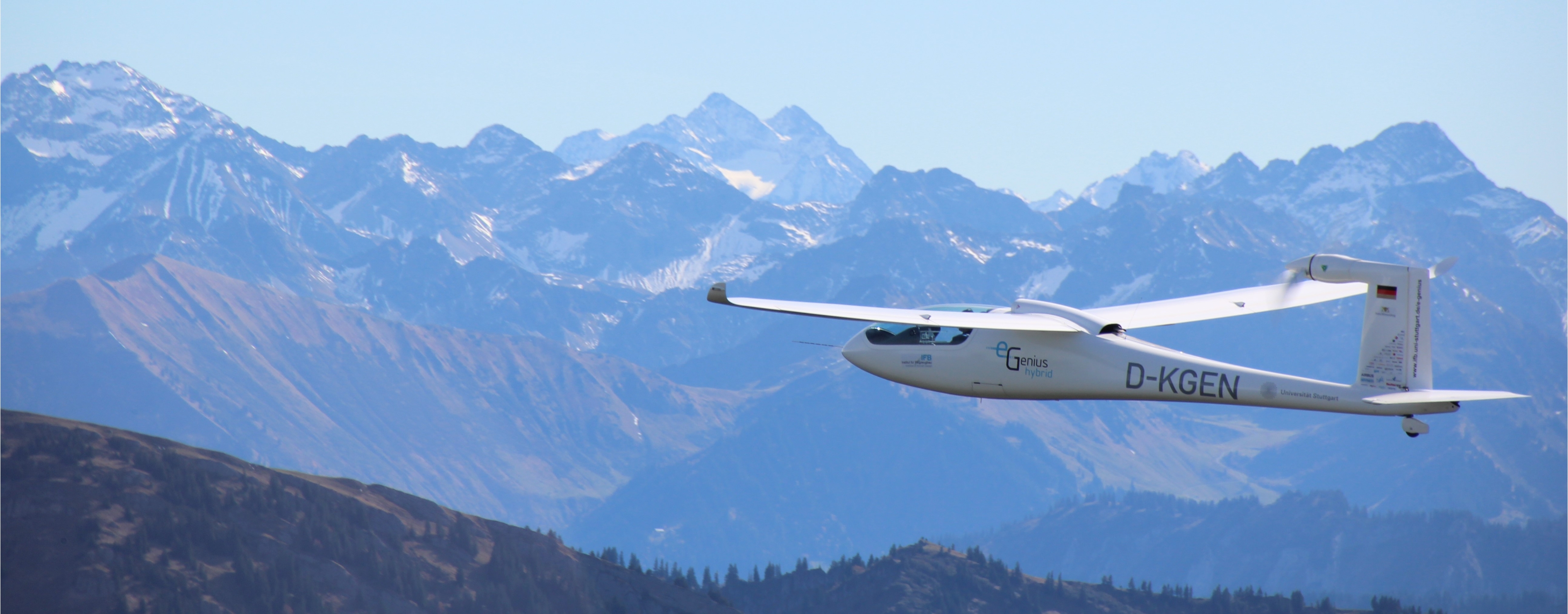 A white electric airplane with the inscription 'e-Genius hybrid' flies in front of an impressive mountain backdrop with snow-covered peaks and green mountain slopes.